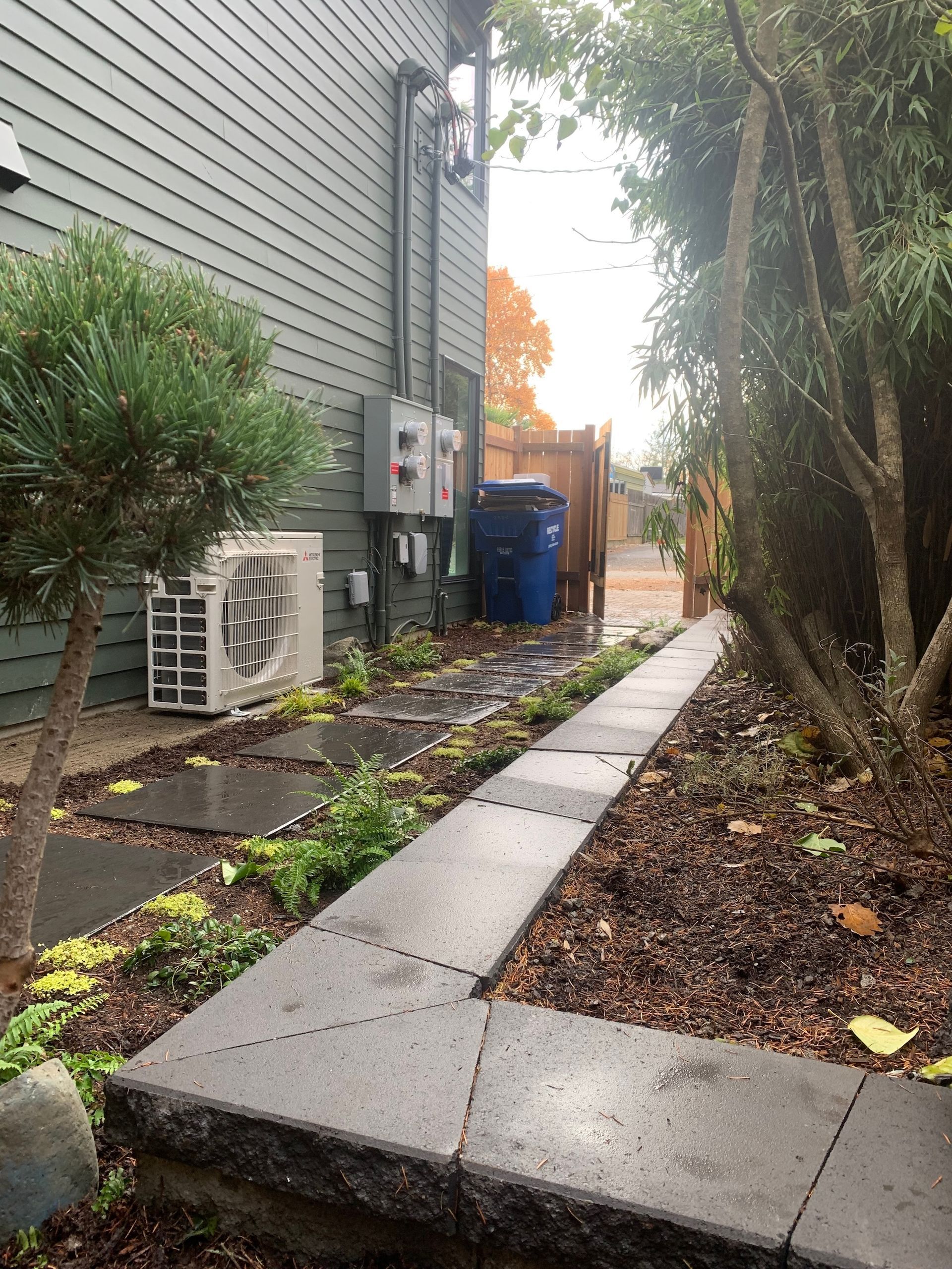 Narrow concrete walkway beside a gray building, lined with trees, leaves, and utility units on a wet day