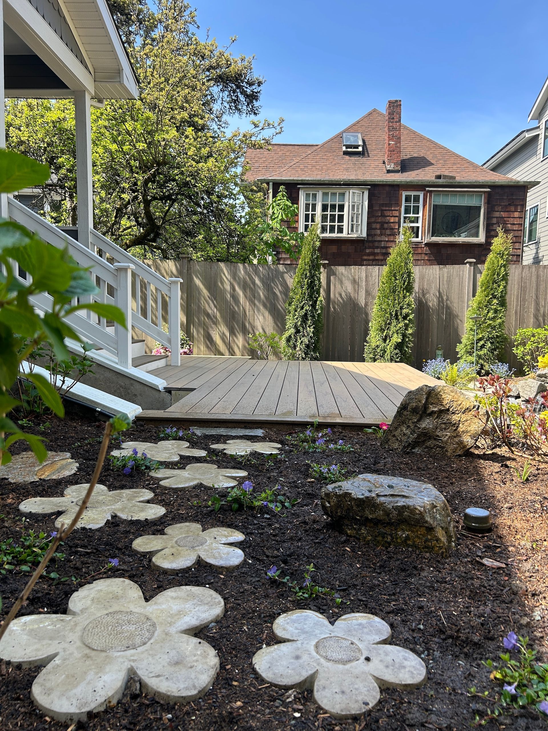 Small backyard garden with flower-shaped stepping stones, wooden deck, fence, and house in the background
