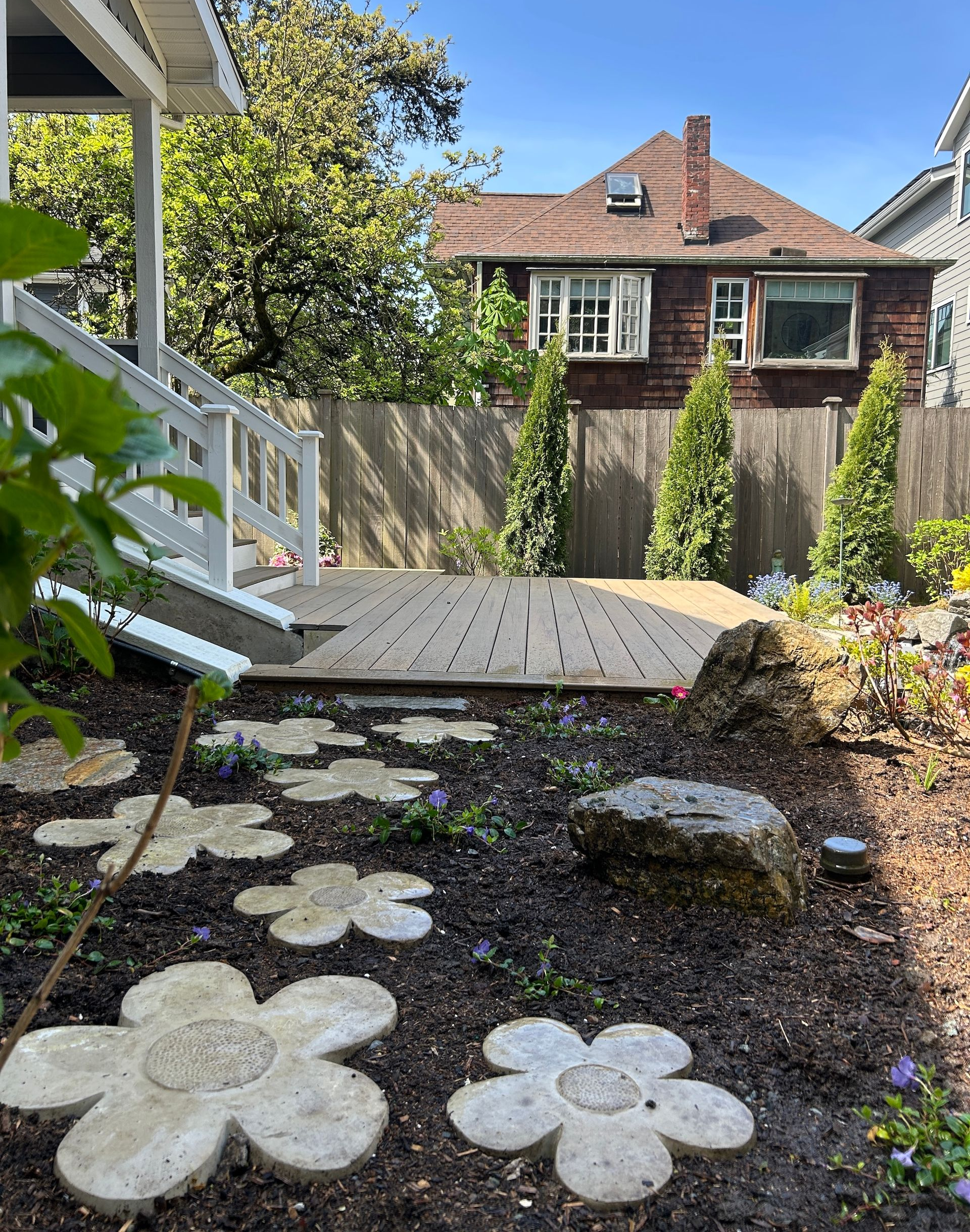 Backyard garden with stone path, wooden deck, flowers, fence, and neighboring house in the background