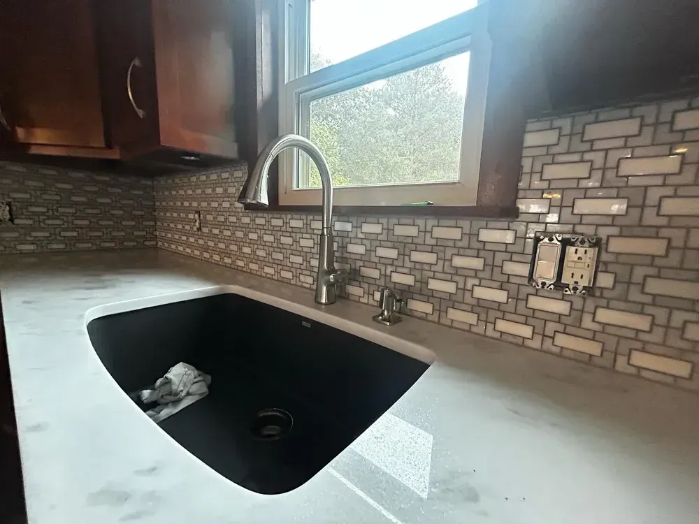 Kitchen sink with black basin, gray countertop, mosaic backsplash, and window above.