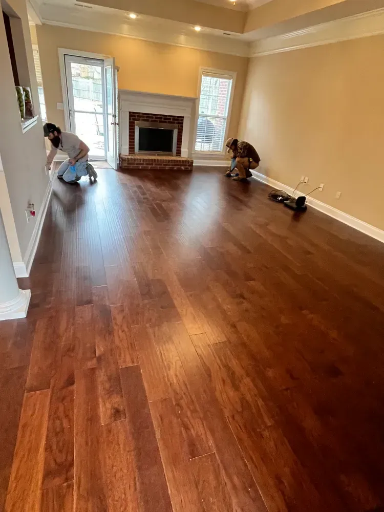 Two people installing dark hardwood flooring in a living room, near a fireplace and french doors.