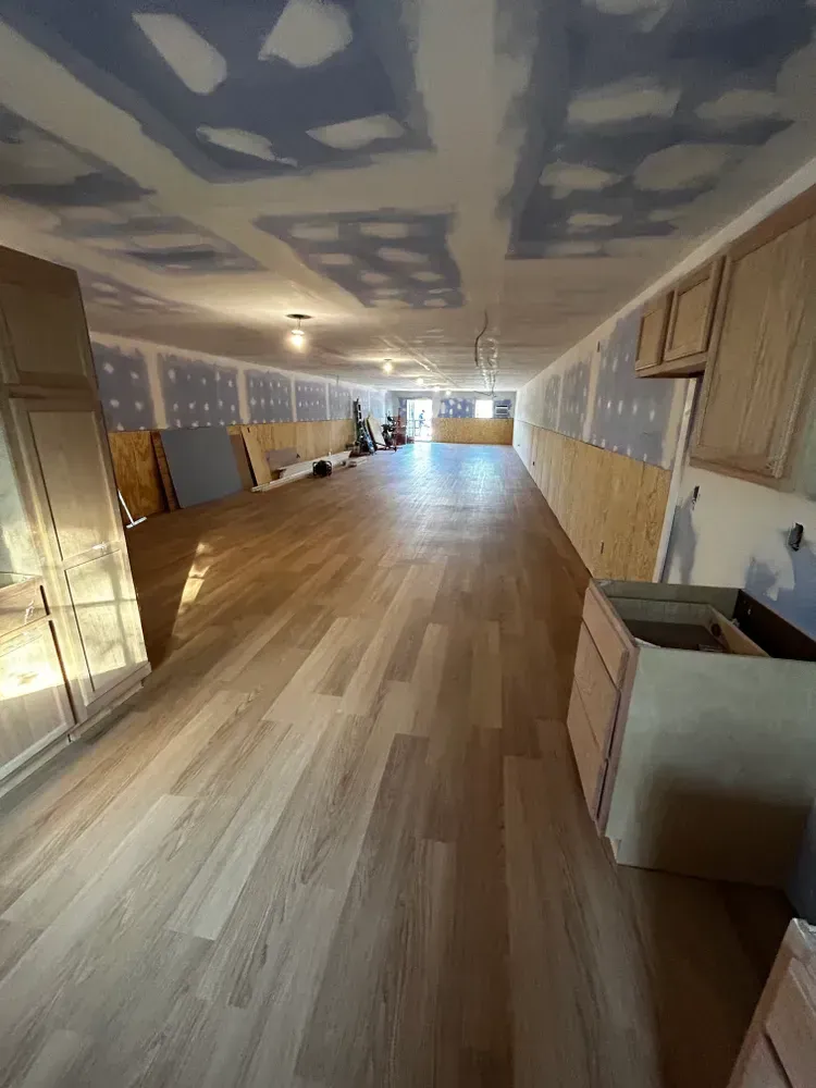 Long, narrow room under construction with new wood-look flooring and exposed drywall.