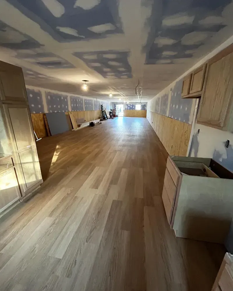 Long, unfinished room with wood floors, drywall, cabinets, and wood paneling on walls.