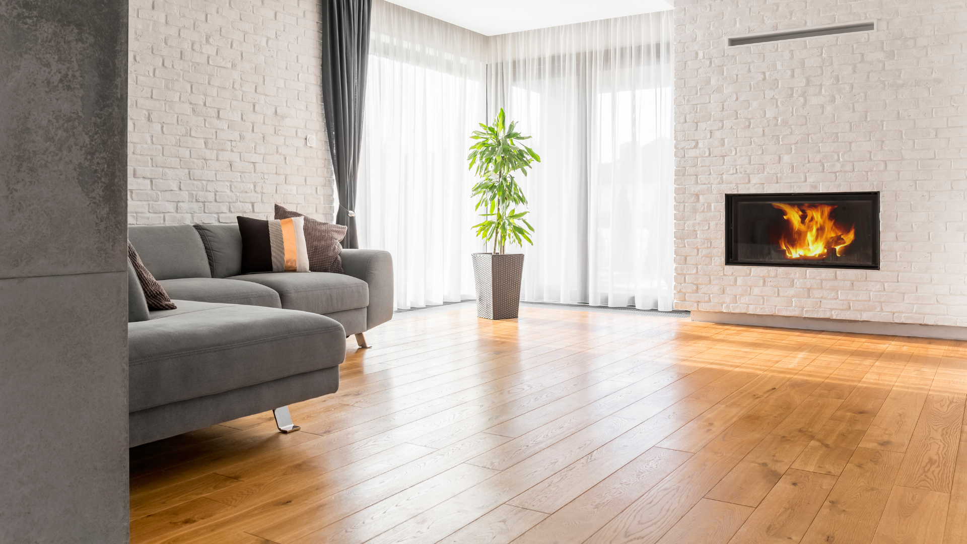 Living room with a gray sectional, fireplace, brick wall, sheer curtains, wood floors, and potted plant.