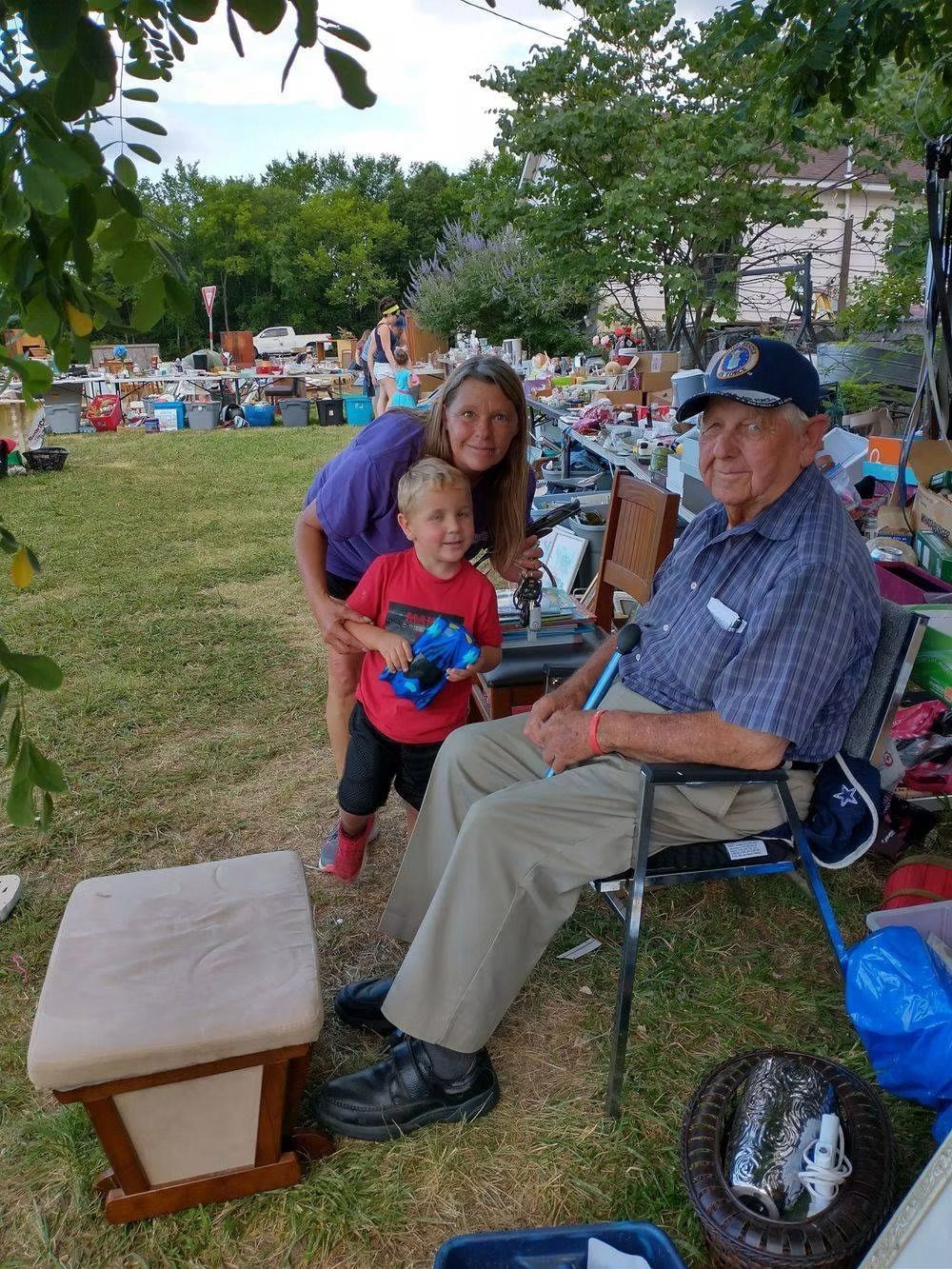 Woman and child with elderly man seated in yard with yard sale items.