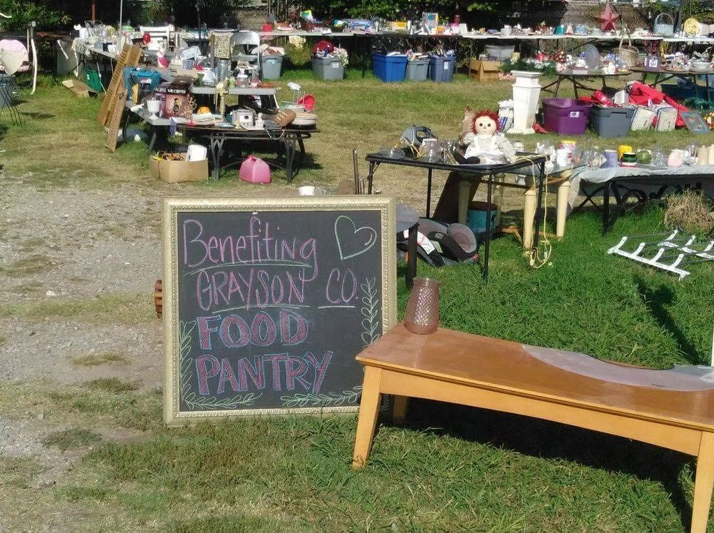 Yard sale benefiting Grayson County Food Pantry with tables of items on grass. Chalkboard sign.