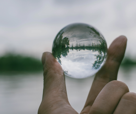 Mano sosteniendo una esfera de cristal transparente que refleja un paisaje de agua y árboles.