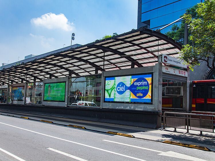 Una estación de autobuses con pantallas publicitarias digitales, una marquesina y una calle en un entorno urbano.