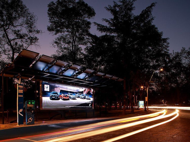 Parada de autobús con paneles solares y pantalla digital publicitaria de coches BMW por la noche. Estelas de luz de los coches que pasan.