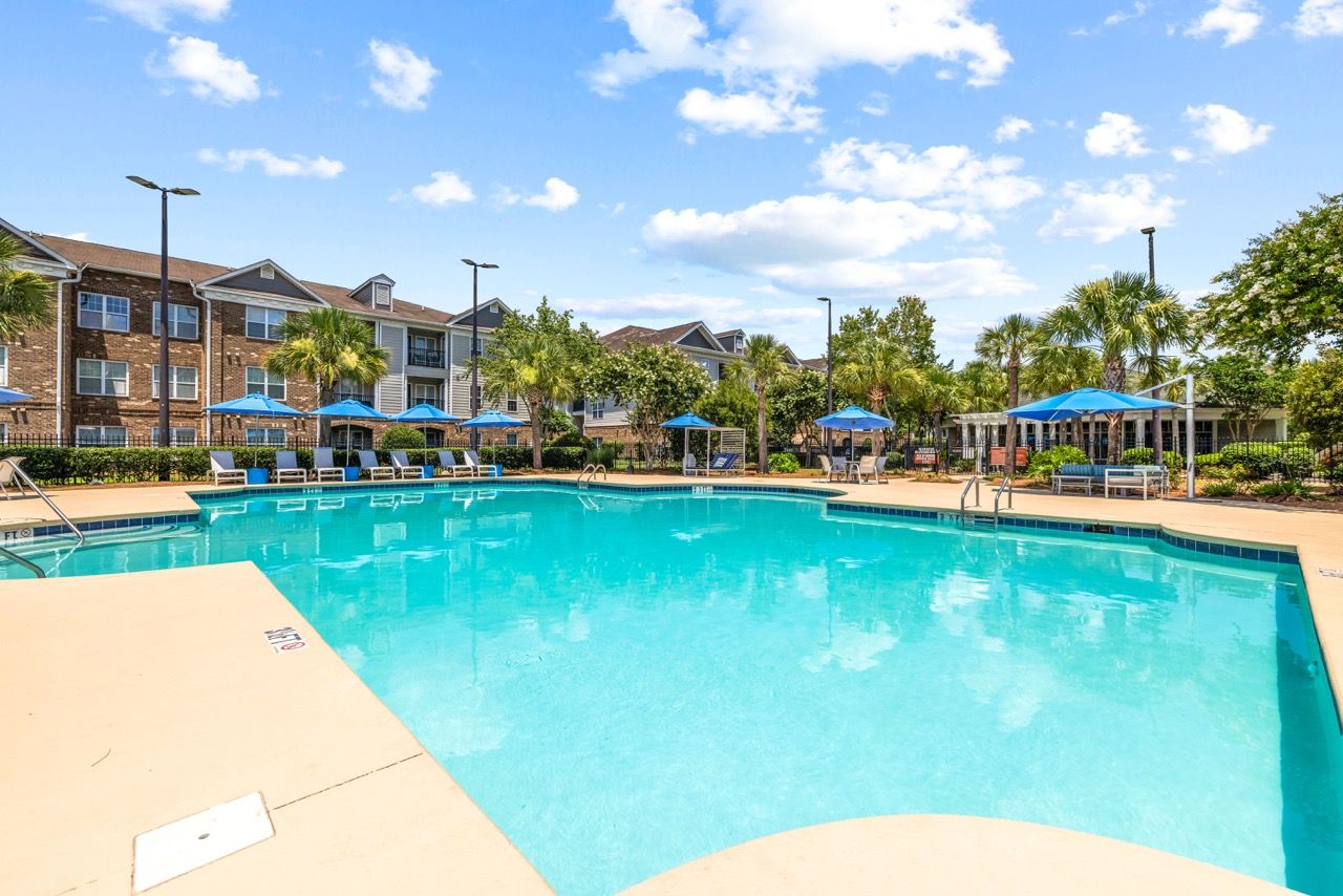 Outdoor swimming pool at a multifamily community with blue umbrellas and palm trees.