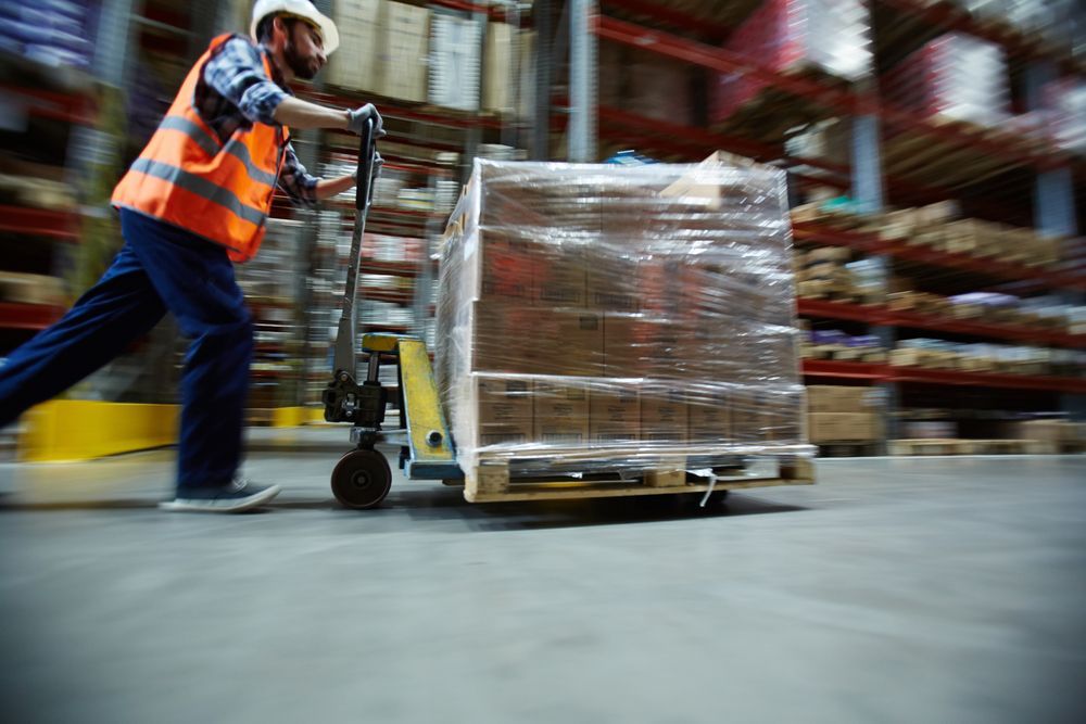 motion shot of warehouse worker wearing hardhat and reflective jacket pushing moving cart with boxes along isle between tall racks