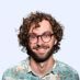 Man with curly brown hair, glasses, smiling, wearing a patterned shirt.