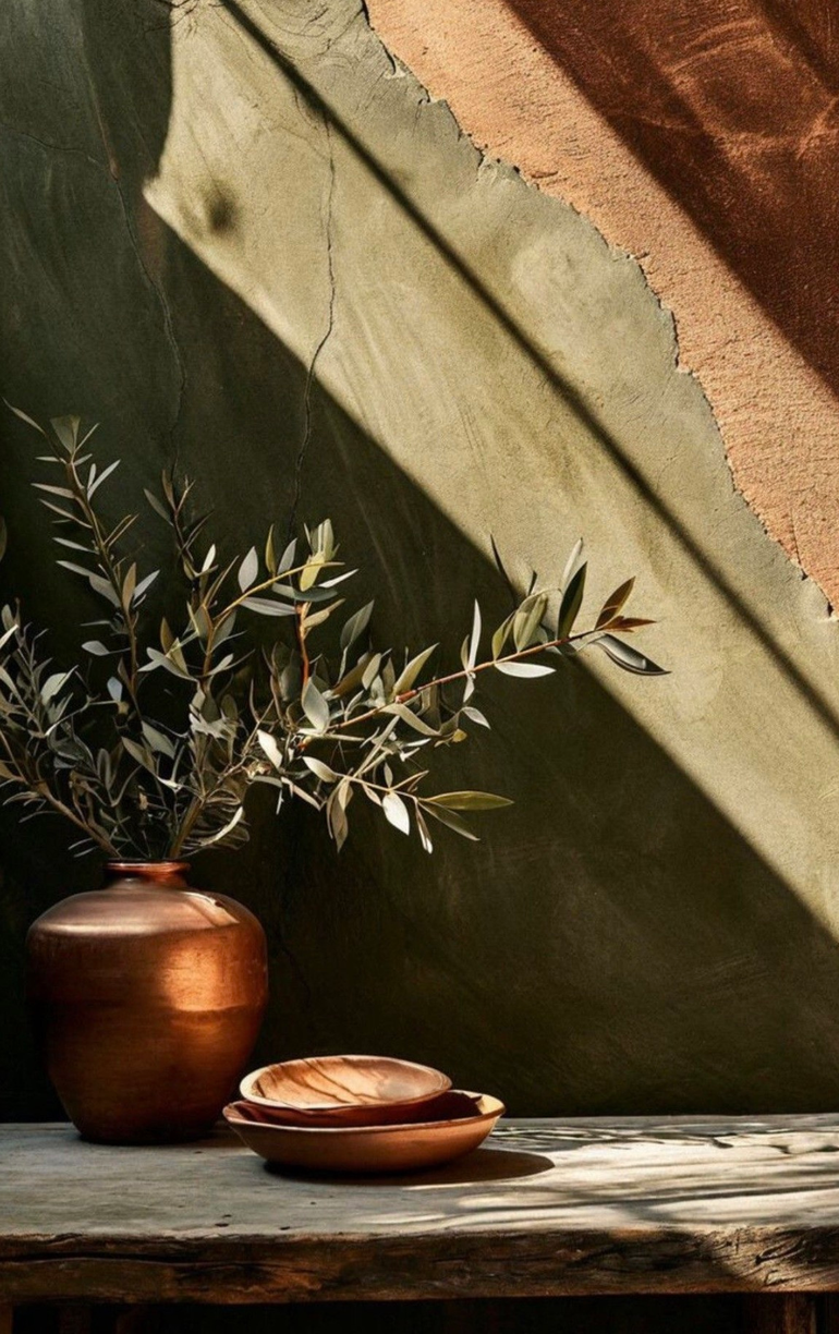 Rustic vase with leafy branches and a bowl on a sunlit wooden table against a green wall.