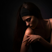 Woman in a dark setting, looking down and touching her shoulder, lit with soft dramatic light