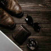 Brown shoes, wallet, sunglasses, and coffee on a dark wooden desk with a laptop corner visible