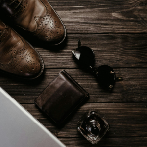 Brown shoes, wallet, sunglasses, and coffee on a dark wooden desk with a laptop corner visible