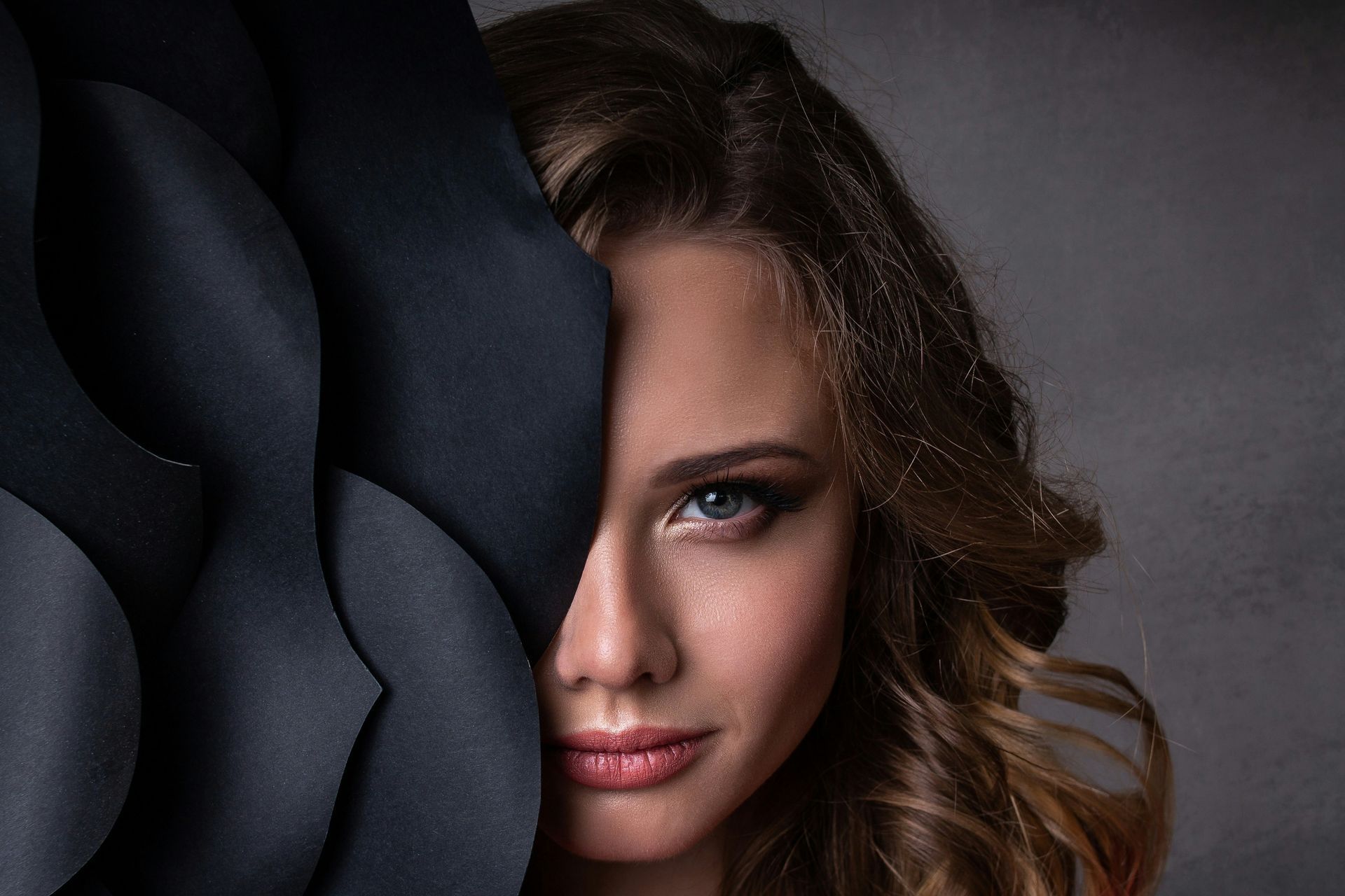 Woman peeking through overlapping dark paper folds, with one eye and part of her face visible