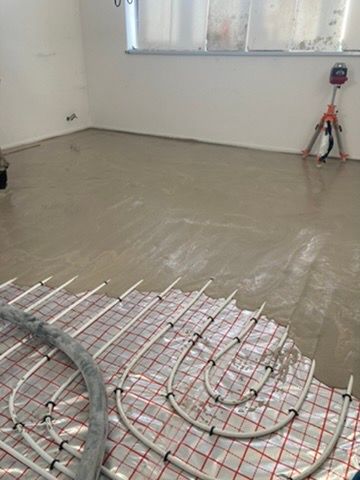 Místnost ve výstavbě s čerstvou betonovou podlahou, odkrytými trubkami sálavého vytápění a oknem v zadní části.