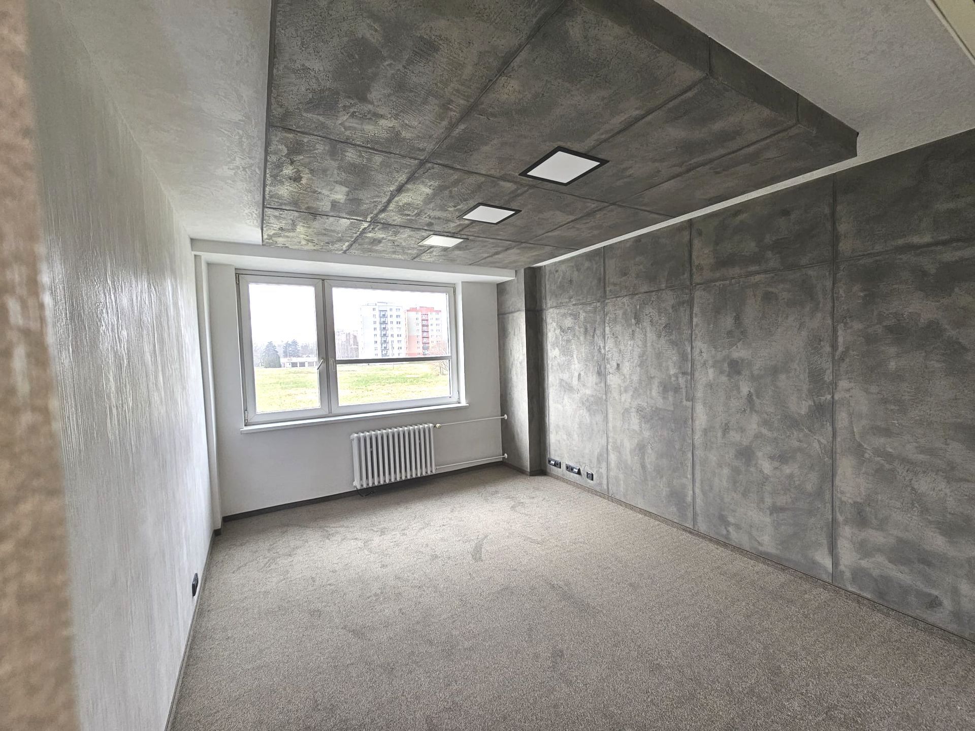 Prázdný moderní pokoj s šedými betonovými stěnami, kobercem a oknem s radiátorem pod ním.