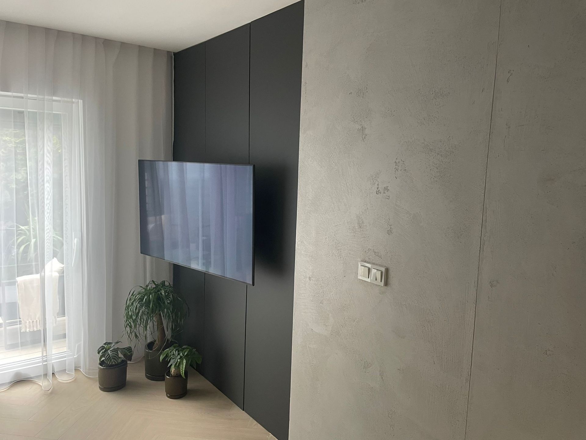 Moderní obývací pokoj s nástěnnou televizí, tmavou akcentovou stěnou, průsvitnými závěsy a květináči u světlého okna