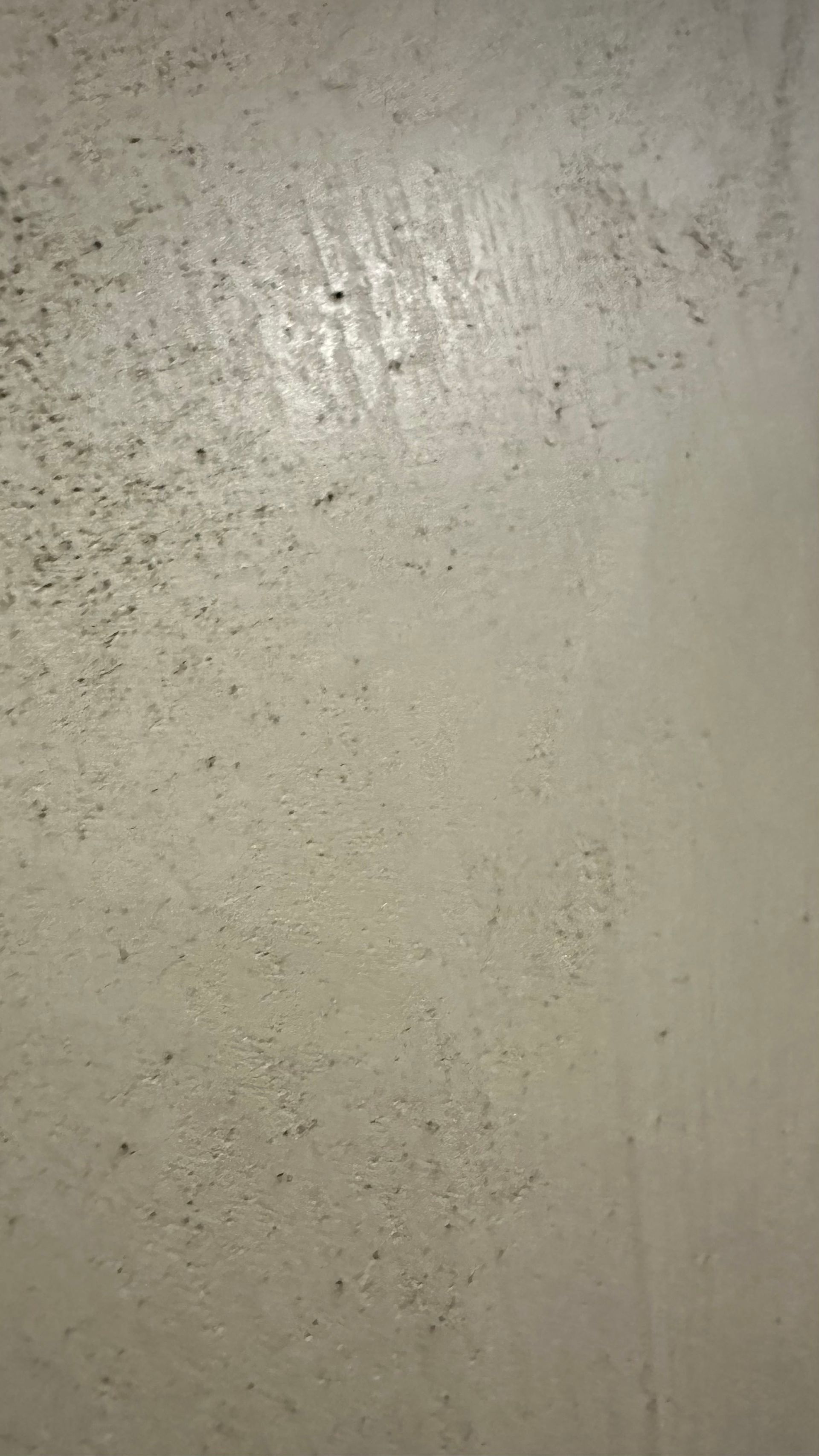 Detailní záběr na béžovou, texturovanou zeď s malými tmavými skvrnami a jasným odrazem světla blízko vrcholu.