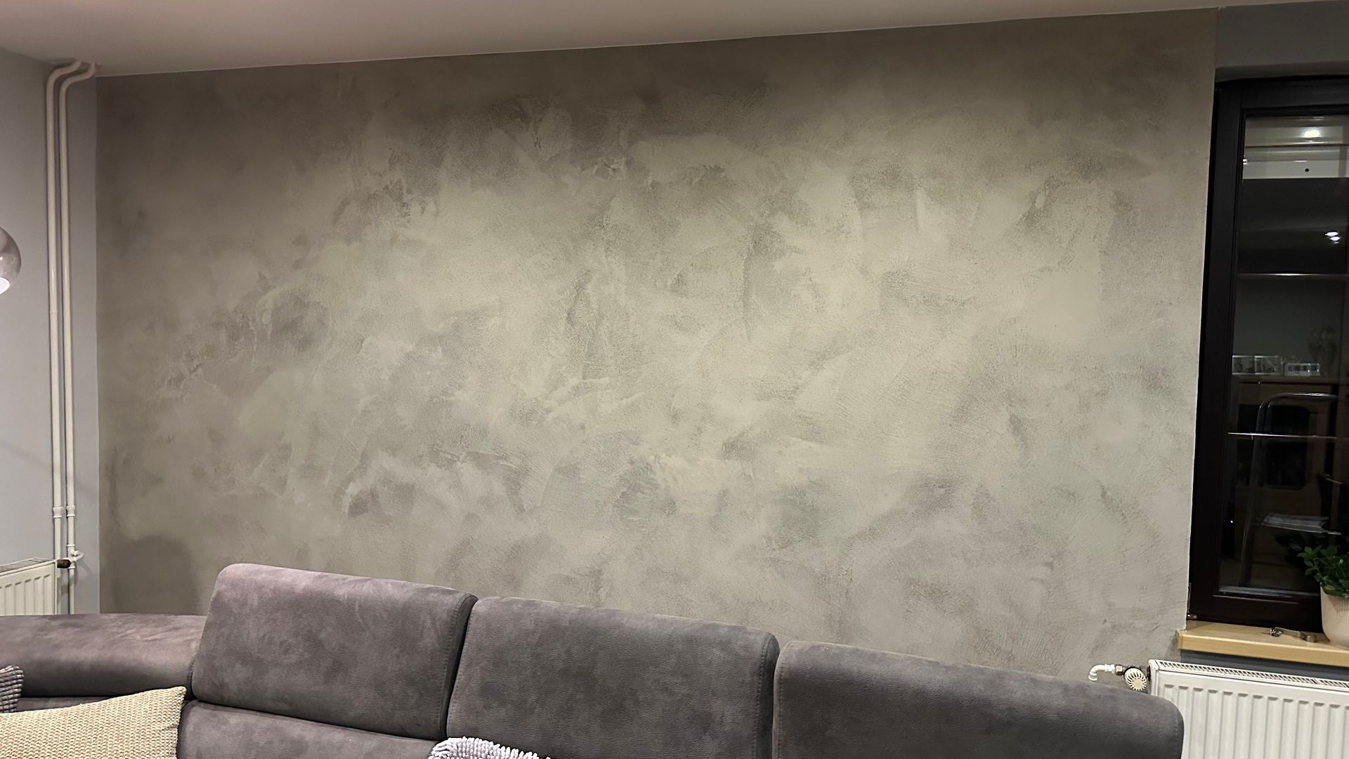 Šedá texturovaná stěna nad tmavou pohovkou v obývacím pokoji s oknem a radiátorem vpravo.