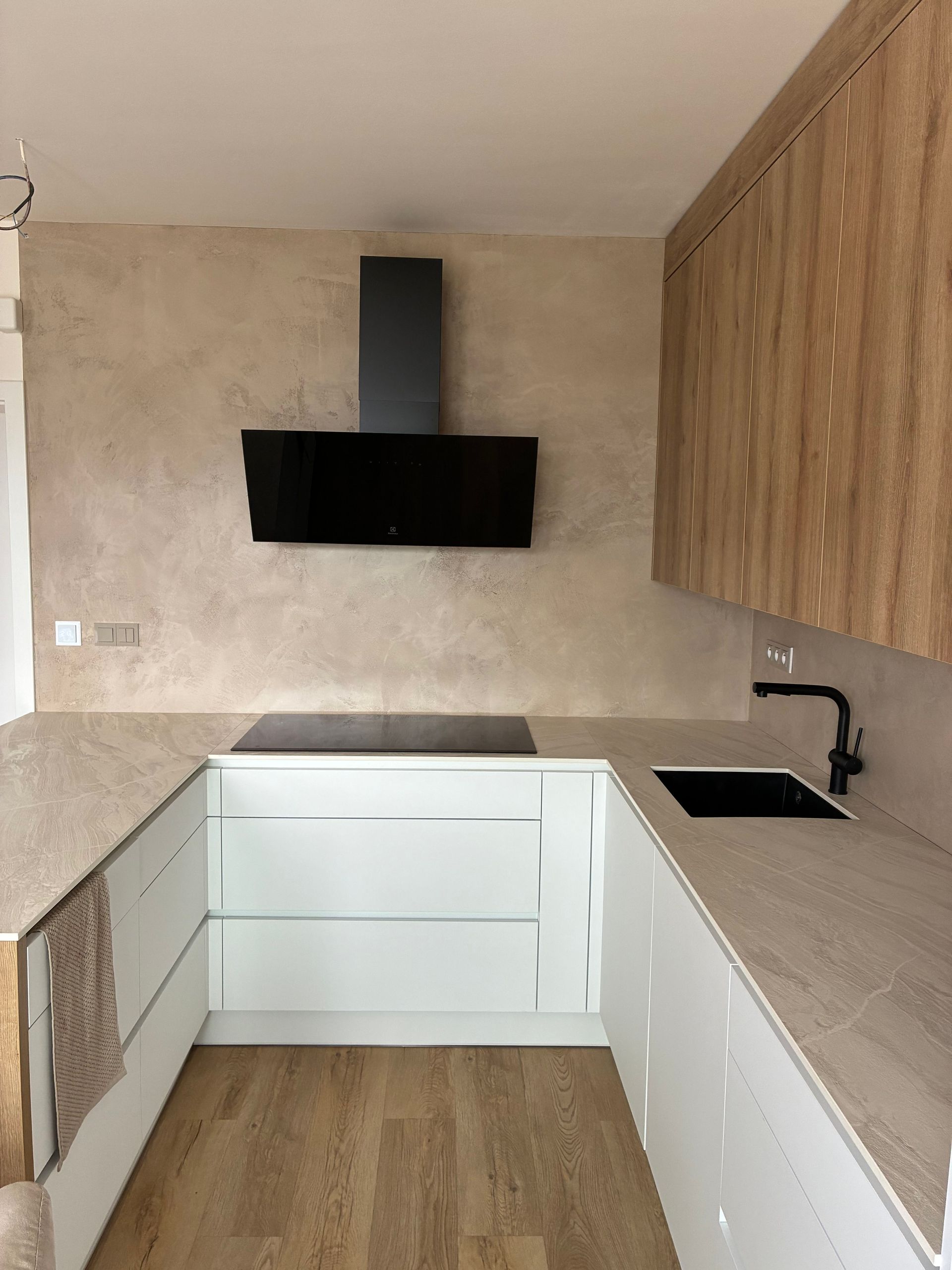 Moderní kuchyně s bílými skříňkami, dřevěnými stěnovými panely, béžovými deskami a černou digestoří.
