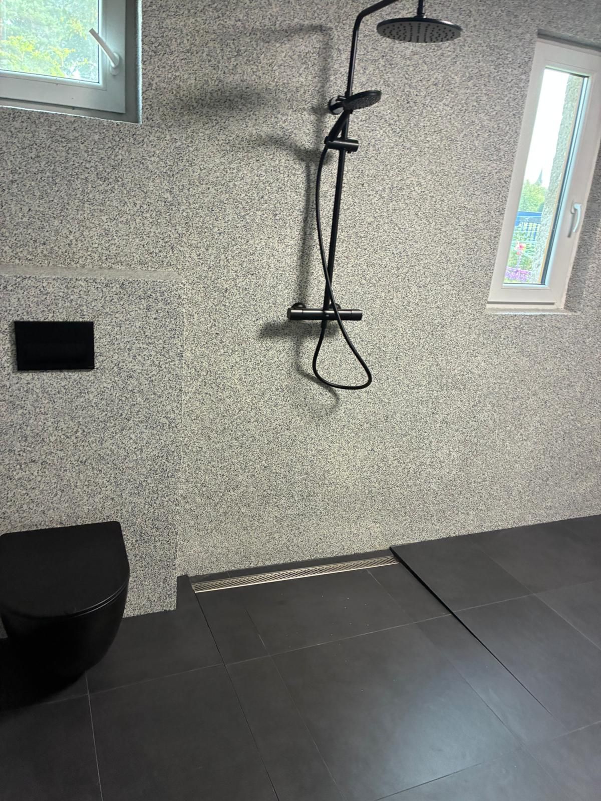 Moderní sprchový kout s tmavou dlaždicovou podlahou, skvrnitými stěnami, černými armaturami a oknem vpravo.
