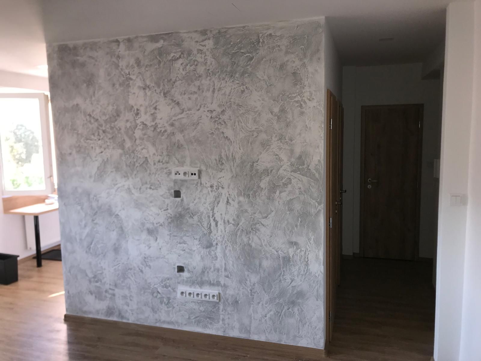 Vnitřní pokoj s šedou texturovanou stěnou, dřevěnou podlahou, dveřmi a oknem vlevo.