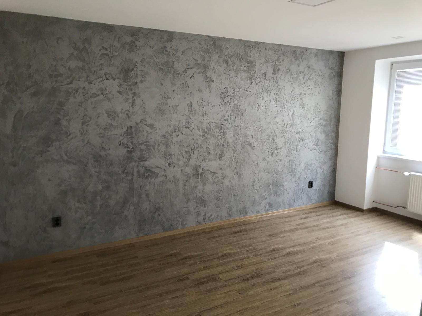 Prázdný moderní pokoj s šedou texturovanou stěnou, dřevěnou podlahou a světlým oknem vpravo.