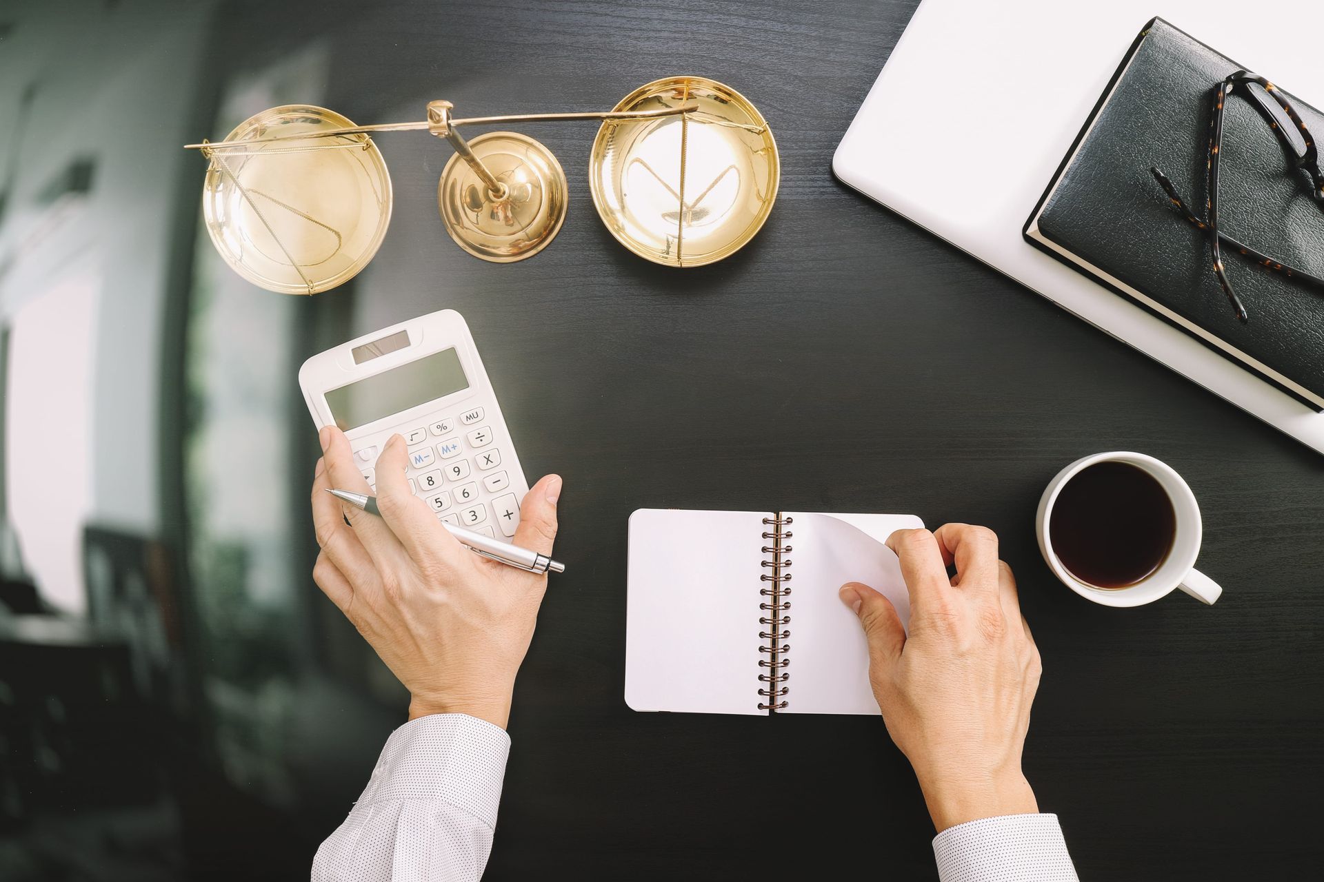 Overhead view of hands using a calculator and notebook on a black desk with gold decor and a coffee cup