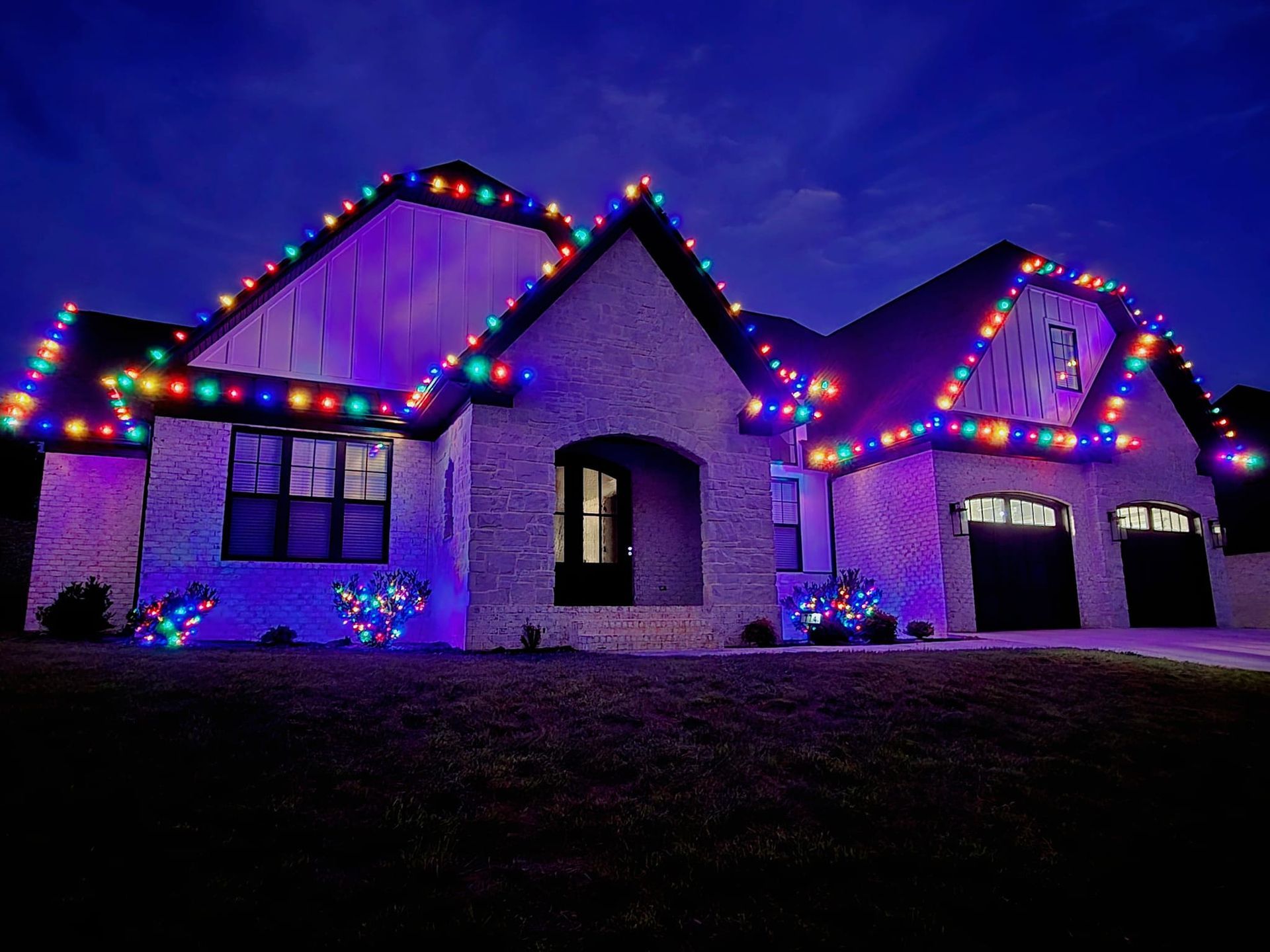 A brick house at night decorated with colorful Christmas lights along the roof and bushes.