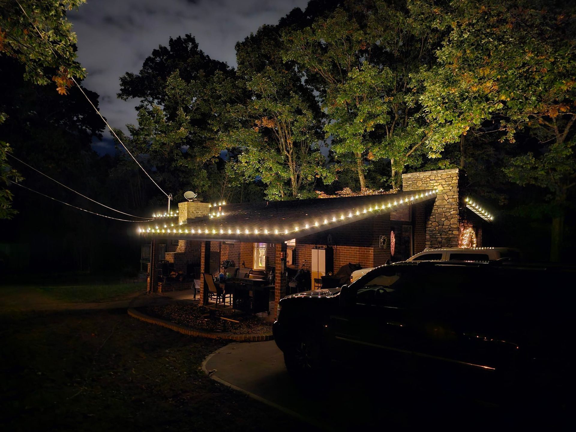 Night view of a cabin with string lights, a car parked in front, trees in the background.
