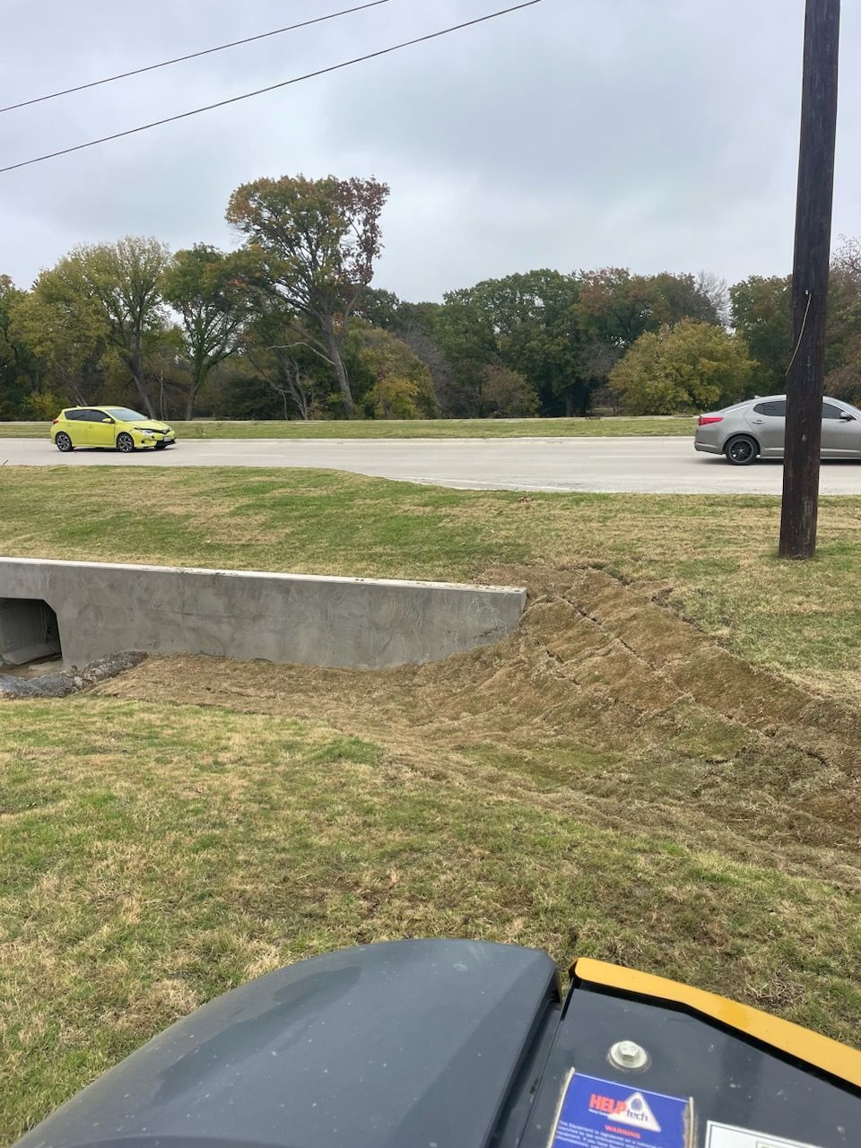 Parking lot with a yellow car, a gray car, grass, and a utility pole near a concrete drain.