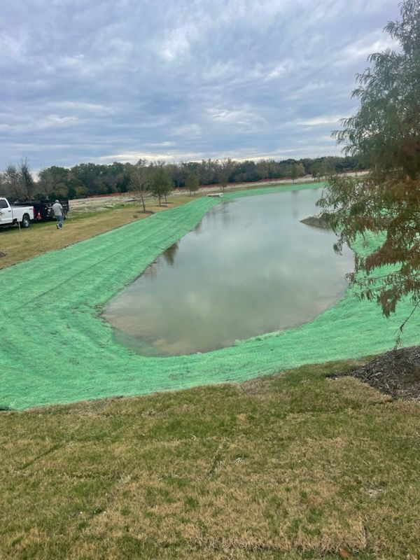 A pond or pond liner covered with green silt fence beside a grassy field under a cloudy sky