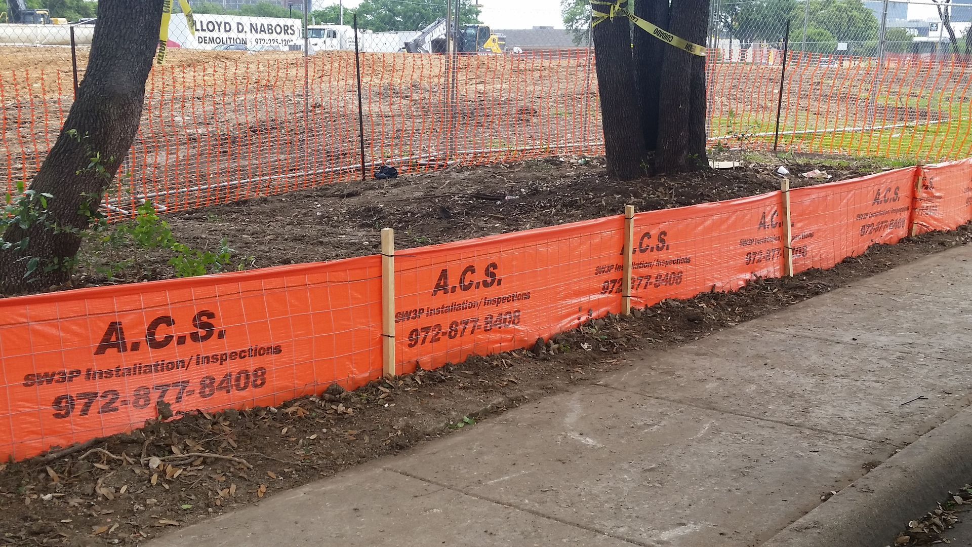 Orange construction fence along a sidewalk beside trees and a dirt lot