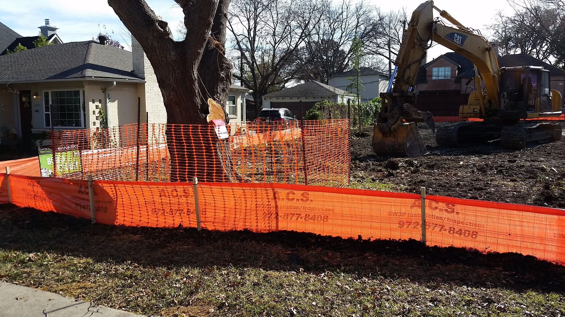 Orange safety fencing around a demolition site with a backhoe and a house in the background