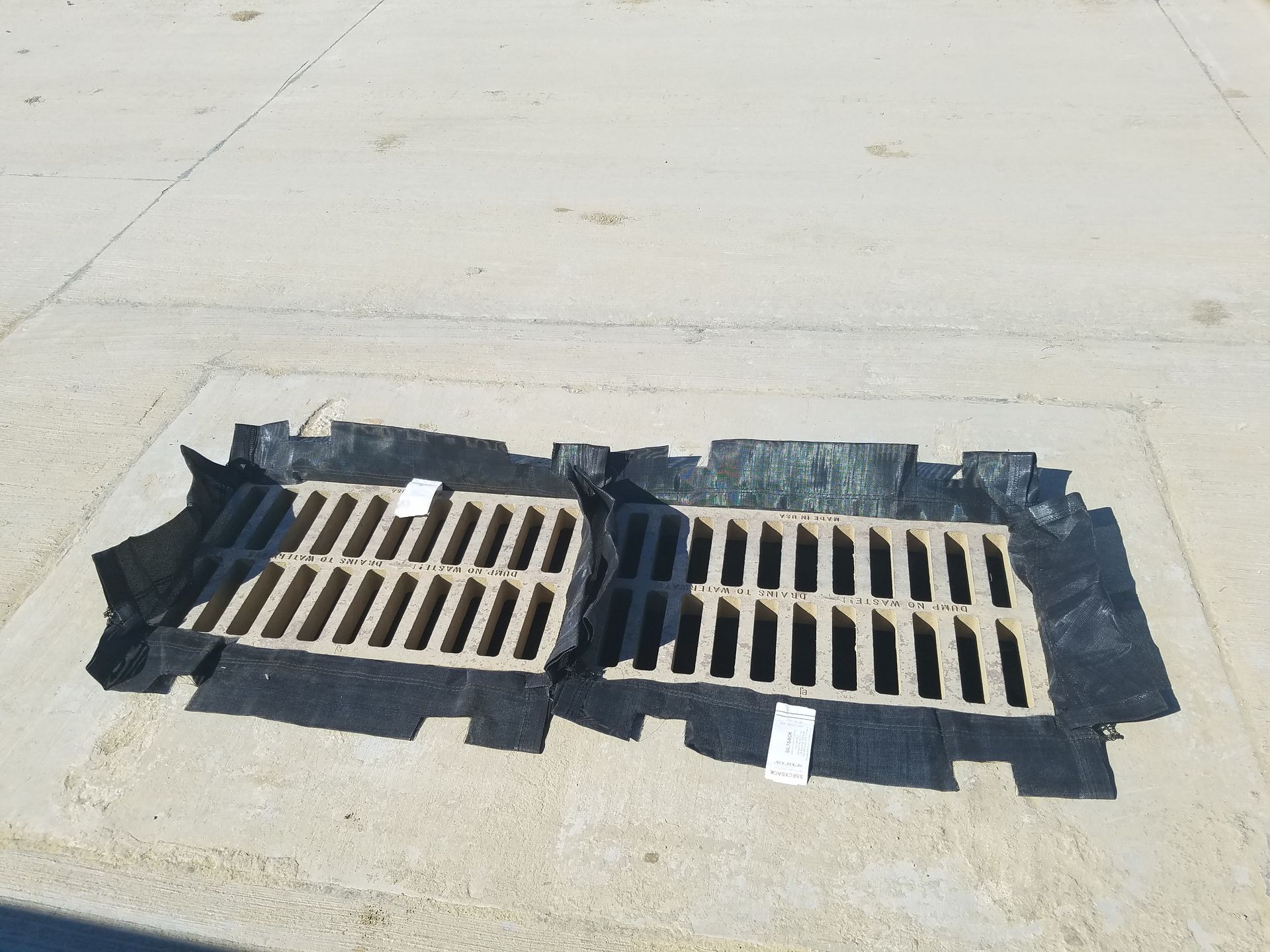 Two metal storm drain grates set in concrete, bordered by black asphalt patches