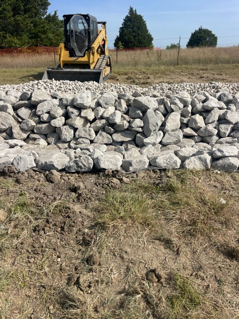 Yellow skid steer behind a pile of gray rocks in a grassy field under a clear sky