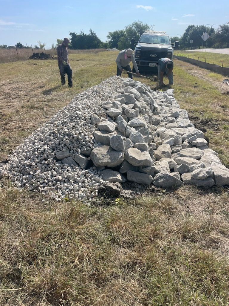 Workers beside a truck and a pile of large rocks on a grassy roadside under a clear sky