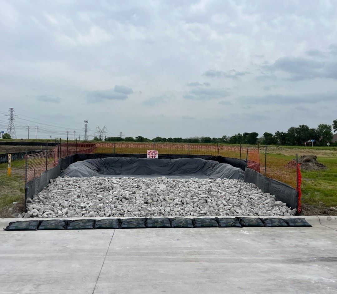Large gravel pit lined with black tarp and orange fencing beside a concrete foreground under cloudy sky