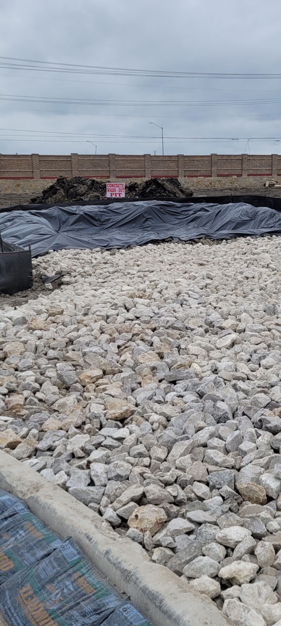 Construction site with gray rocks, black tarp, and a brick wall under a cloudy sky