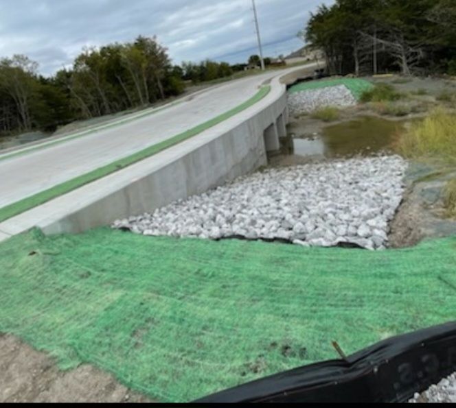 Concrete road embankment under construction with green erosion control mesh and white riprap along a ditch