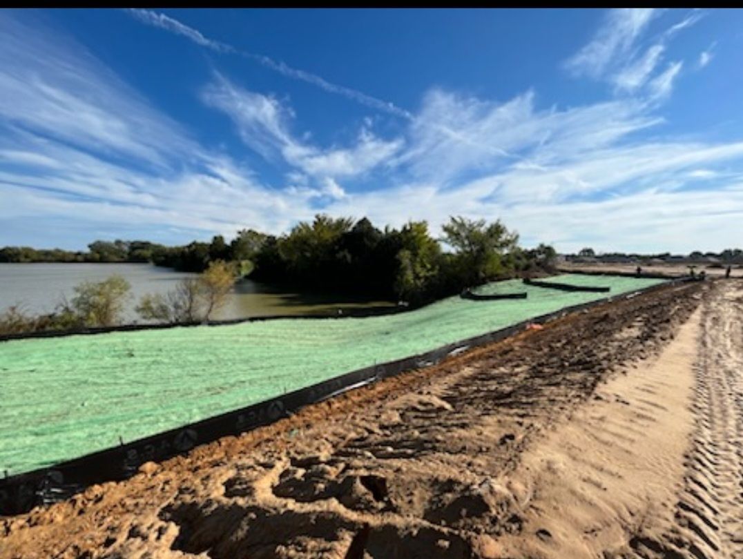 Green erosion-control mats line a muddy dirt road beside a river under a blue sky