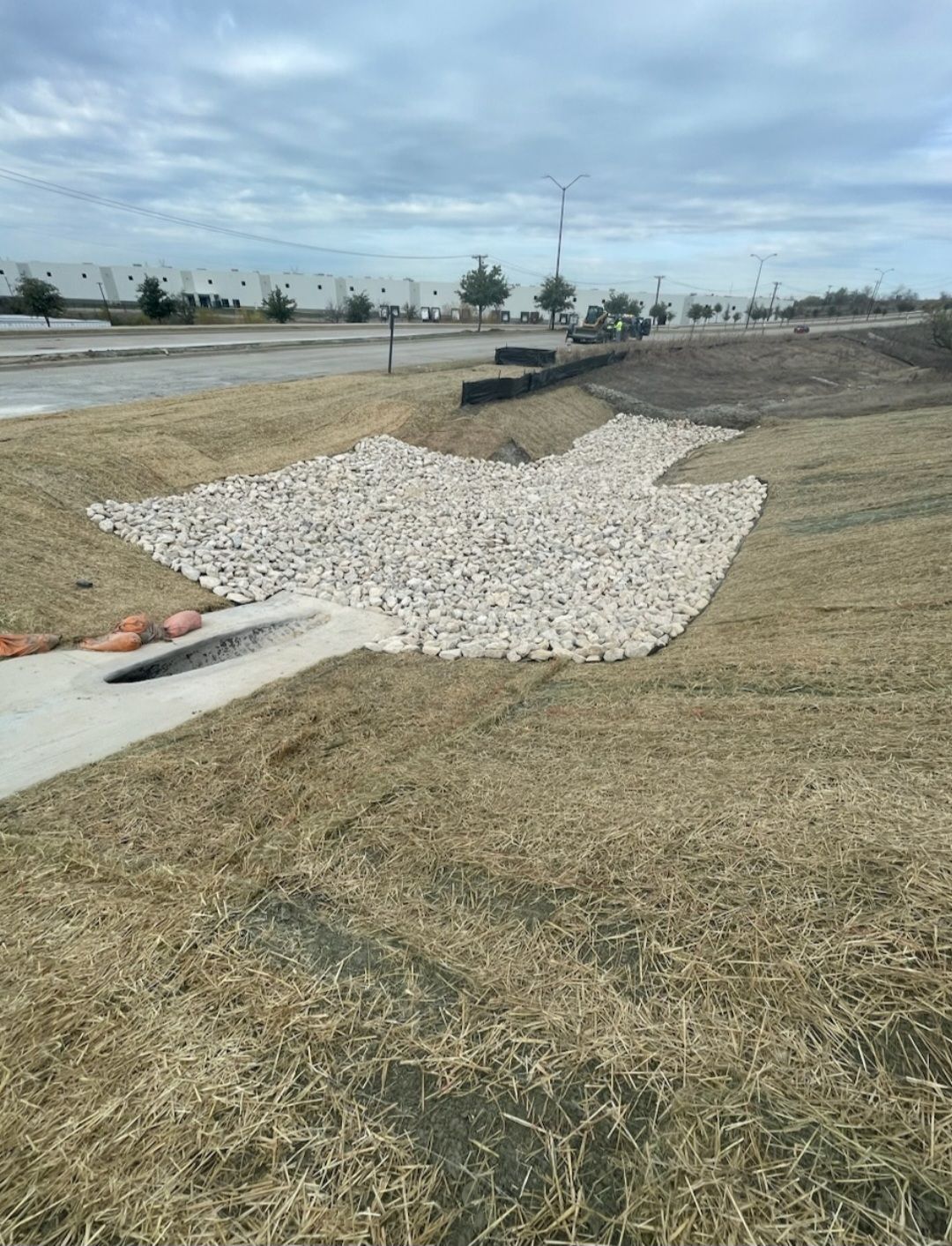 Rock-lined drainage ditch in a grassy roadside area under a cloudy sky.