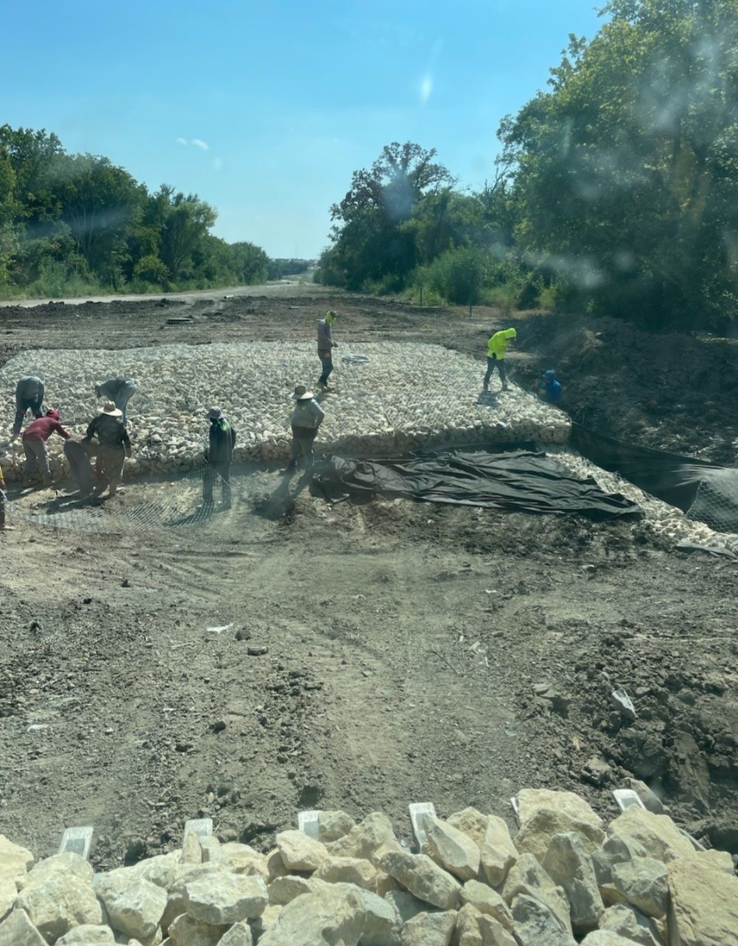 Workers spreading gravel on a rural dirt road beside trees and a ditch