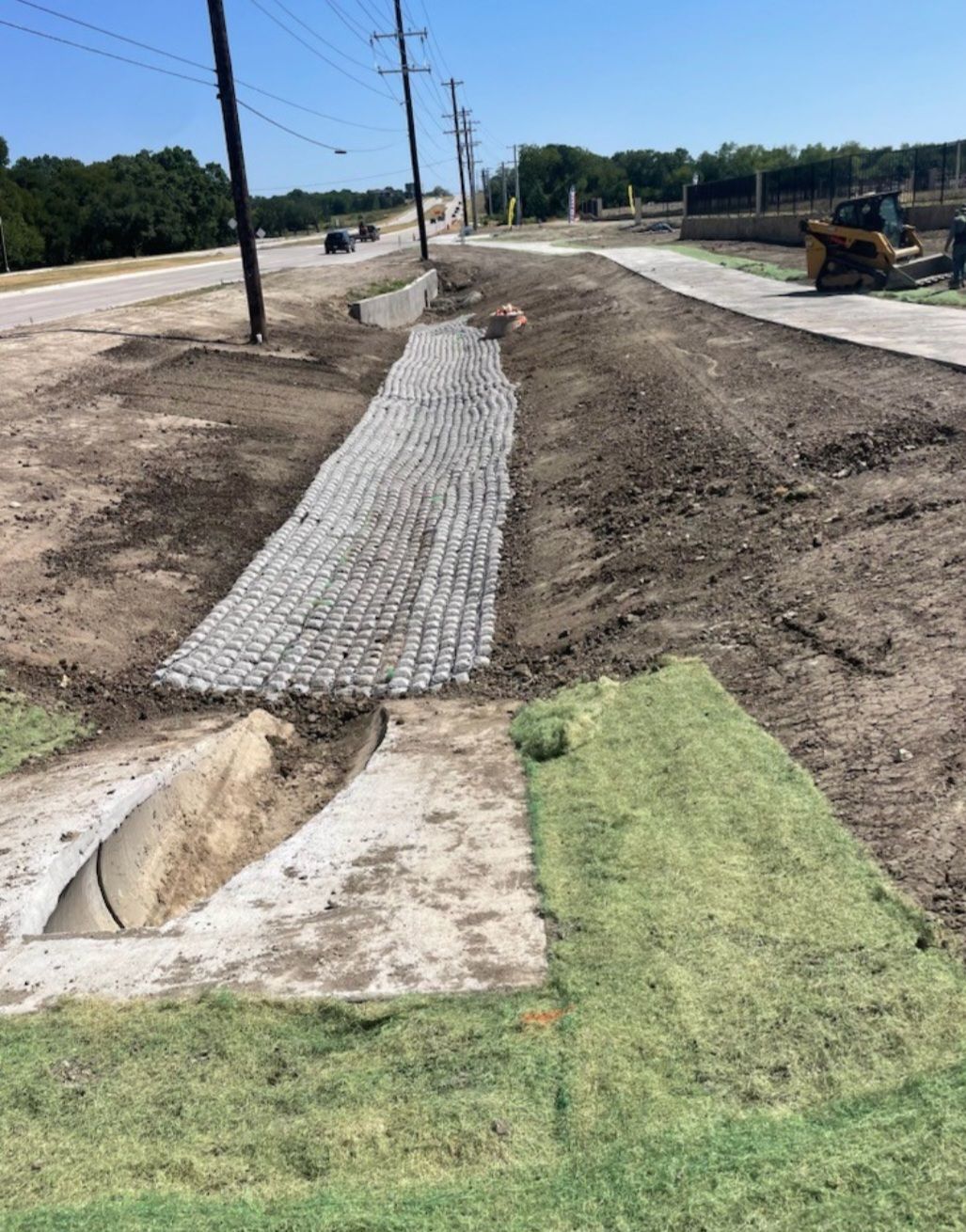 Construction site with a dirt path and concrete drainage channel beside a road under repair