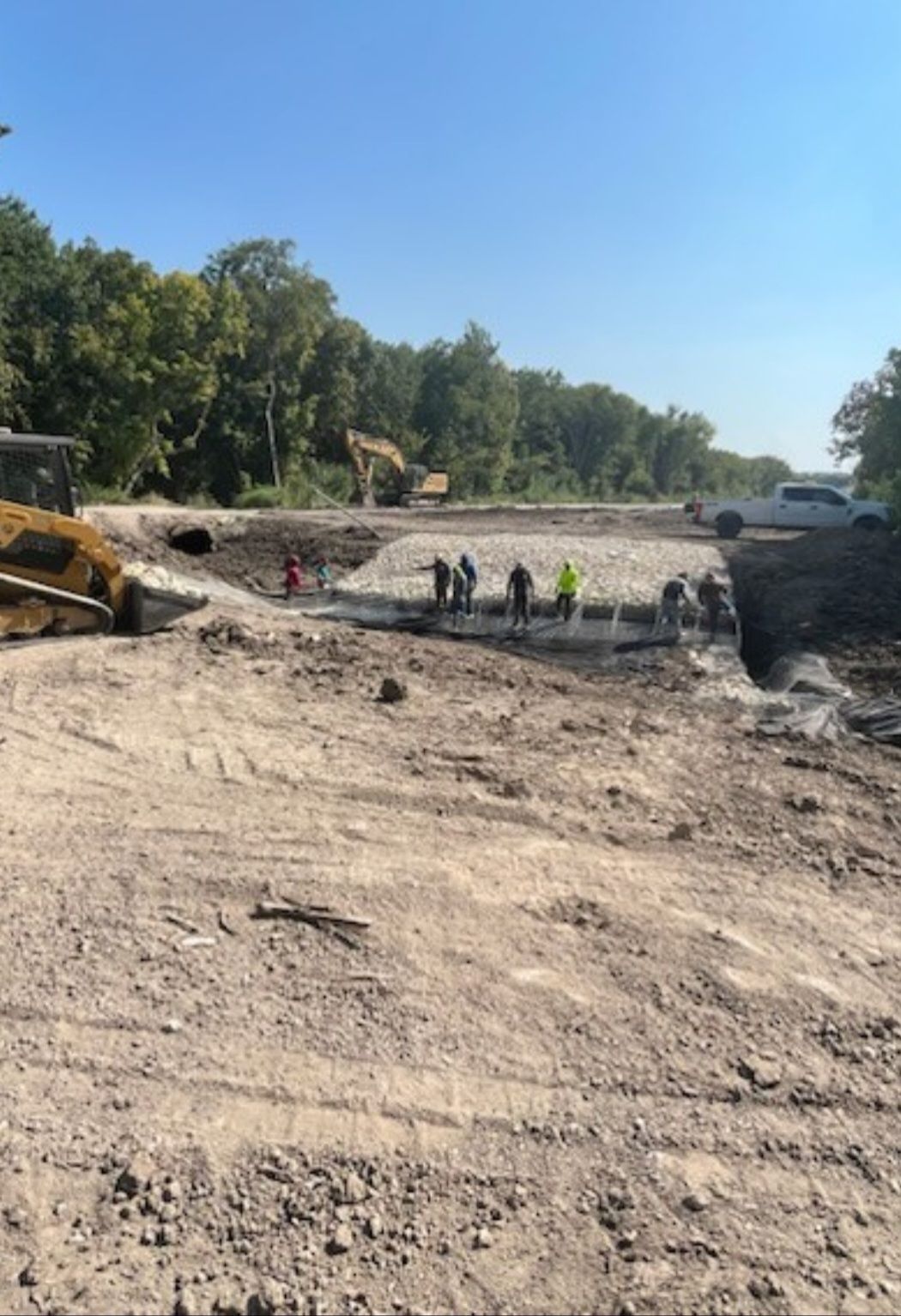 Construction workers excavating a large dirt site with an excavator and trees in the background