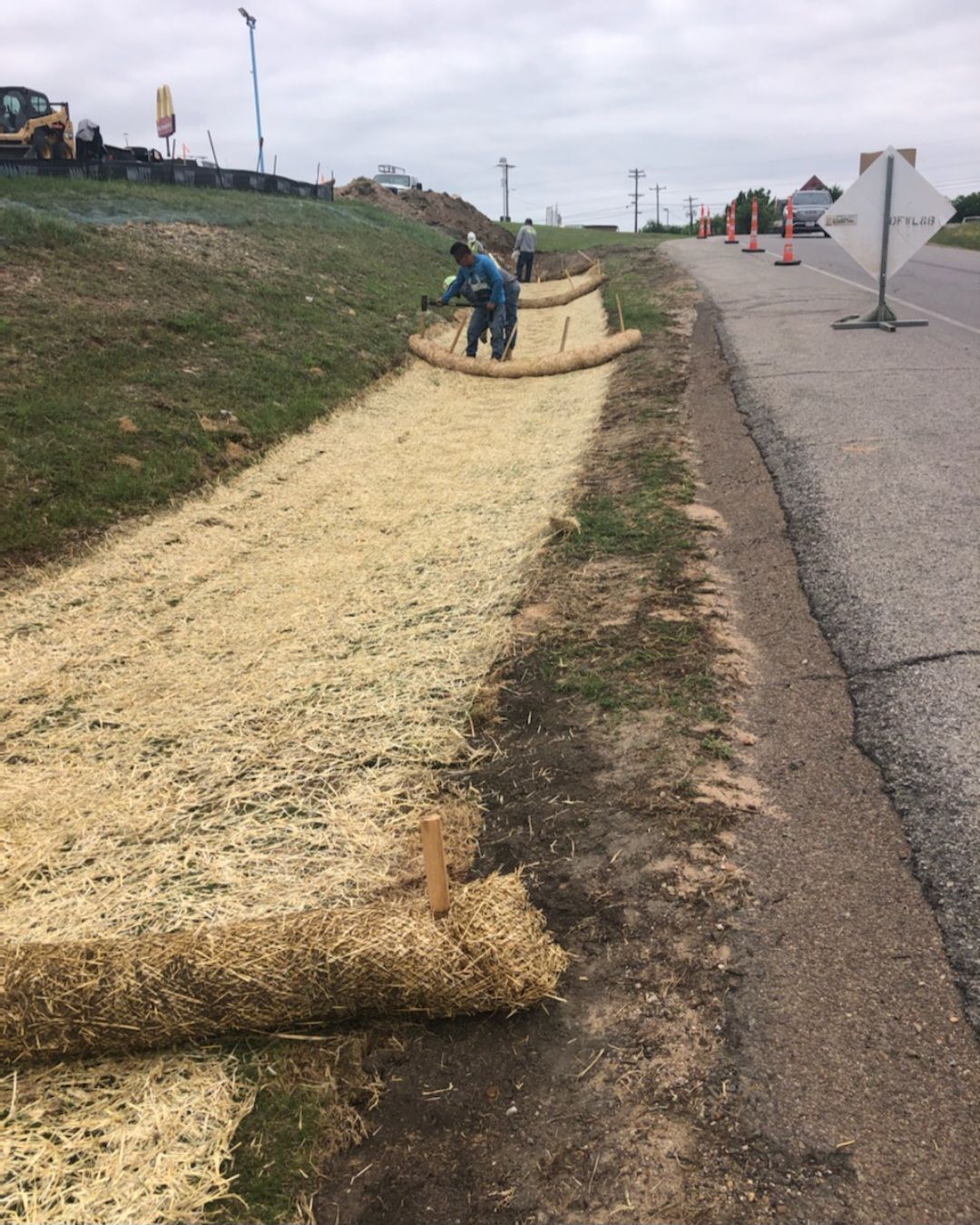 Workers laying straw matting along a roadside embankment under cloudy skies.