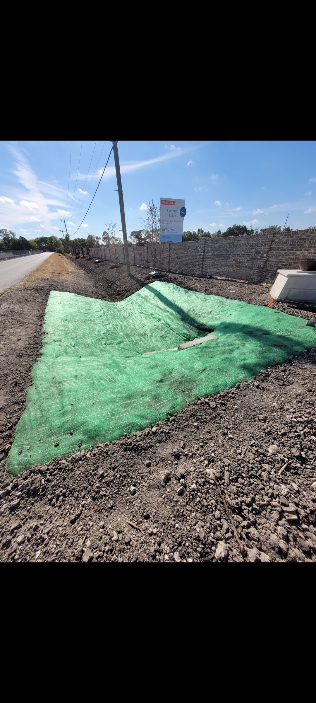 Gravel lot with green tarp-covered mound beside utility poles and a water tower under a clear sky